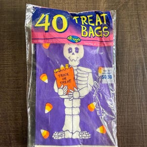 以下が含まれることがあります： Trick or Treatと書かれたオレンジ色の袋を持つ白い骸骨のグラフィックが描かれた、40枚入りの紫色のハロウィーン用トリートバッグの密封パッケージ。パッケージには黄色の文字で40 Treat Bags、Fun Worldのロゴ、キャンディコーンのイラストが印刷されています。