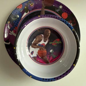 May include: A round dinner plate and a matching bowl featuring Space Jam graphics. The bowl shows a basketball player in a white jersey holding a basketball, with the Space Jam logo and cartoon characters. The plate has a dark purple background.