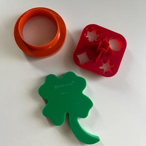 Puede incluir: Tres cortadores de galletas de plástico en naranja, rojo y verde. El cortador naranja es un anillo. El cortador rojo es cuadrado con recortes de estrella, diamante y círculo. El cortador verde tiene forma de trébol. La palabra "Tupperware" es visible.