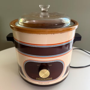 Puede incluir: Una olla de cocción lenta Crock-Pot vintage con exterior en color crema y marrón oscuro, decorada con rayas horizontales naranjas y azules. Incluye una tapa de cristal transparente con pomo, asas laterales negras y un mando central de control.