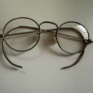Puede incluir: Gafas redondas vintage con montura de metal plateado. Las lentes son transparentes y las patillas se curvan hacia abajo. Las gafas están sobre un fondo blanco liso. Un diseño de gafas clásico.