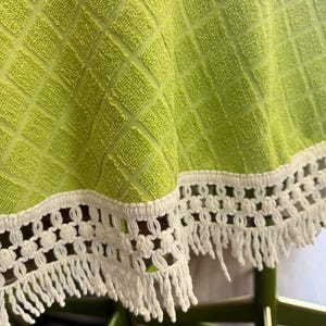 May include: A close-up of a lime green tablecloth with a diamond pattern. The tablecloth has a white crocheted trim with fringe. The tablecloth is on a green table.