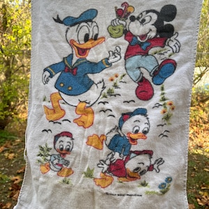 Op de afbeelding: Witte katoenen handdoek met de stripfiguren Donald Duck, Mickey Mouse en Kwik, Kwek en Kwak. De handdoek heeft een gefranjerde rand en is versierd met kleurrijke illustraties van de personages in verschillende poses. De handdoek hangt aan een waslijn.