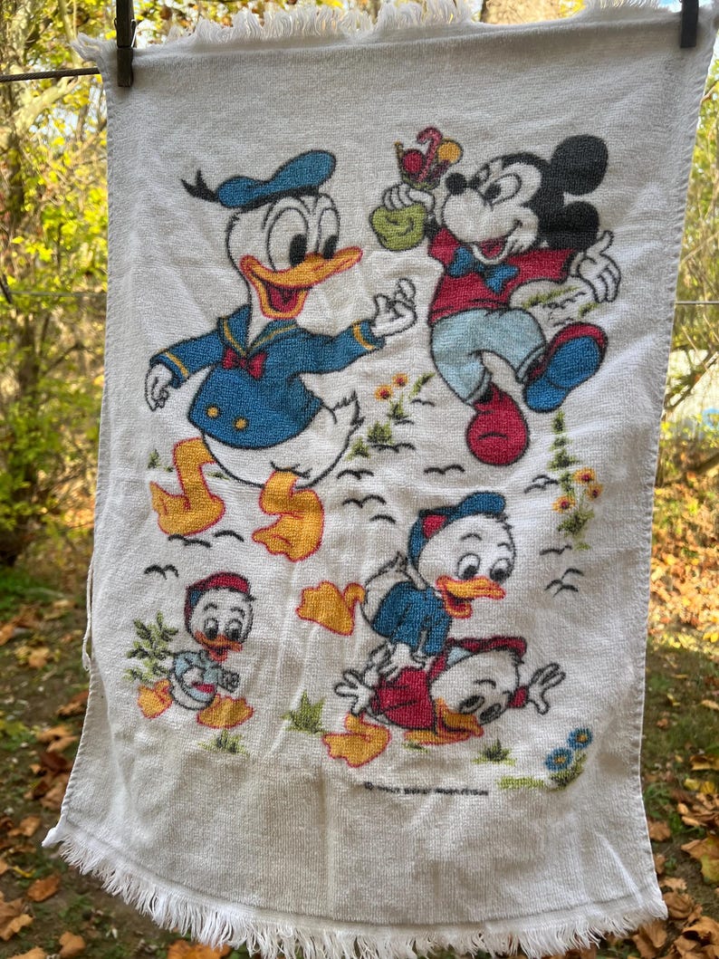 Op de afbeelding: Witte katoenen handdoek met de stripfiguren Donald Duck, Mickey Mouse en Kwik, Kwek en Kwak. De handdoek heeft een gefranjerde rand en is versierd met kleurrijke illustraties van de personages in verschillende poses. De handdoek hangt aan een waslijn.
