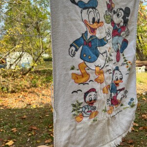Op de afbeelding: Witte vintage handdoek met geborduurde afbeeldingen van Donald Duck en Mickey Mouse. De handdoek heeft een gefranjerde rand en hangt aan een waslijn. De personages zijn afgebeeld in hun klassieke outfits.