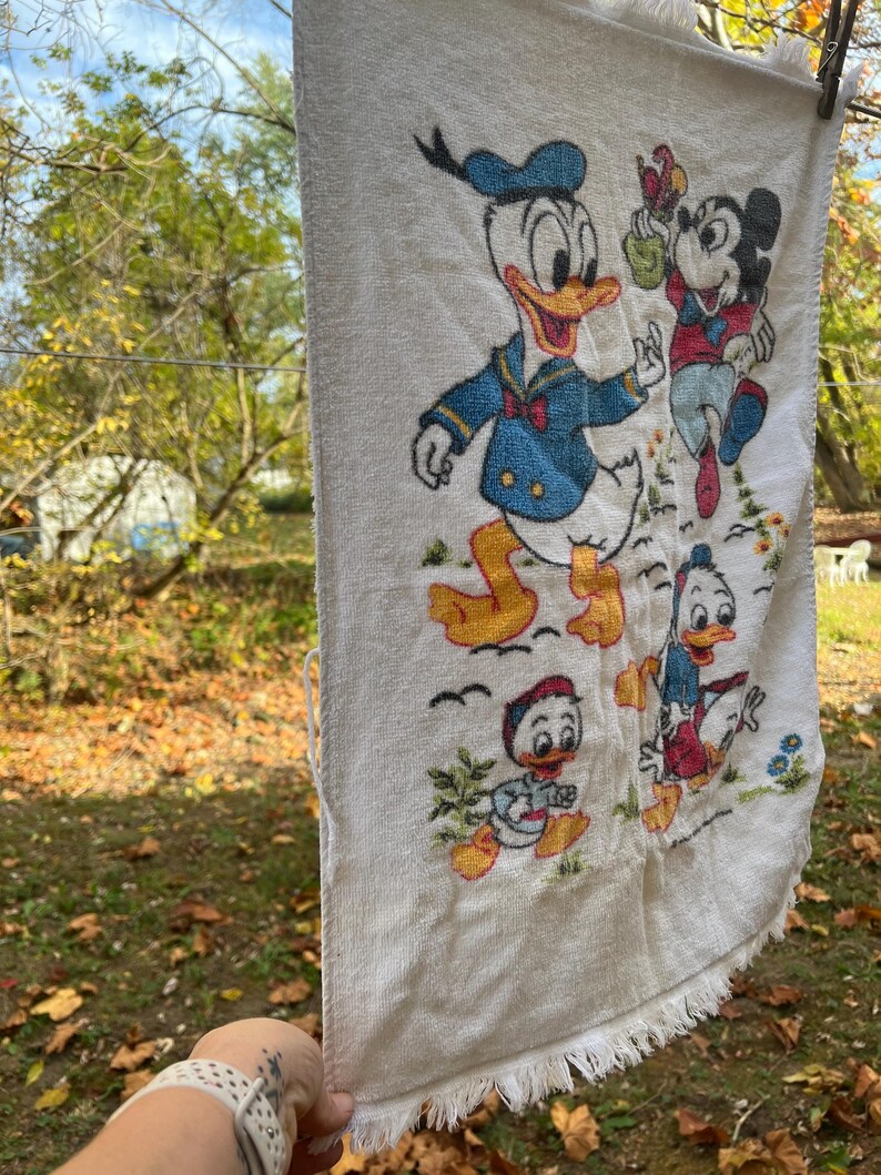 Op de afbeelding: Witte vintage handdoek met geborduurde afbeeldingen van Donald Duck en Mickey Mouse. De handdoek heeft een gefranjerde rand en hangt aan een waslijn. De personages zijn afgebeeld in hun klassieke outfits.
