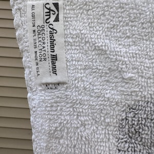 Op de afbeelding: Close-up van een witte badstof handdoek met een label met de tekst "Fashion Manor Decorator Collection". De handdoek is gemaakt van volledig katoen en is gemaakt in de VS. De handdoek heeft een getextureerd oppervlak.