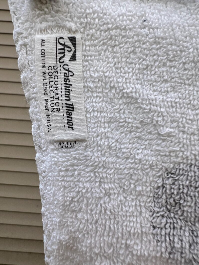 Op de afbeelding: Close-up van een witte badstof handdoek met een label met de tekst "Fashion Manor Decorator Collection". De handdoek is gemaakt van volledig katoen en is gemaakt in de VS. De handdoek heeft een getextureerd oppervlak.