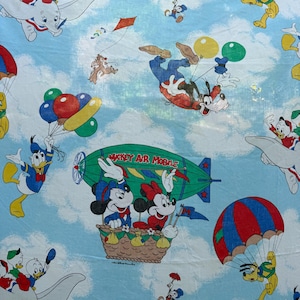 Puede incluir: Tela azul claro con varios personajes de Disney volando. Mickey y Minnie Mouse están en un globo aerostático, mientras que el Pato Donald, Goofy y otros se ven con globos, paracaídas y cometas. El texto "Mickey Air Mobile" es visible.