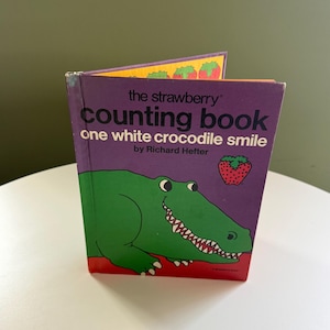 Puede incluir: Un libro infantil vintage titulado "The Strawberry Counting Book" de Richard Hefter. La portada es morada con una ilustración de cocodrilo verde y una fresa. El libro está abierto, revelando páginas coloridas.
