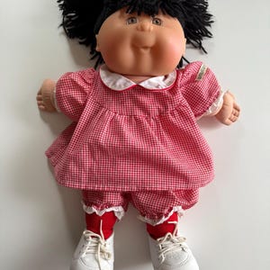 May include: A Cabbage Patch Kid doll with black yarn hair in pigtails wearing a red and white gingham dress and white shoes.