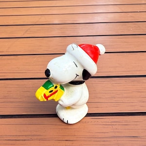 May include: A Snoopy figurine wearing a red Santa hat and holding a yellow and green object. The figurine is white with black accents and a black collar. The figurine is on a brown wooden surface.