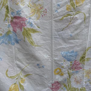 Puede incluir: Tela blanca con un patrón floral con flores rosas, azules, amarillas y verdes. La tela tiene una textura sutil y una apariencia ligeramente arrugada.