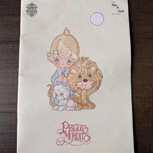 May include: A cross-stitch pattern booklet titled Peace on Earth Book PM-4 by Designs by Gloria & Pat. The cover features a Precious Moments illustration of a child with blonde hair, a small white lamb, and a lion, all rendered in a grid pattern.
