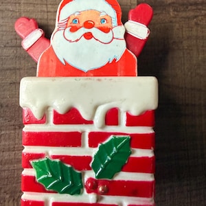 May include: A decorative Santa Claus figure emerging from a red and white brick chimney. Santa has a white beard, red hat, and red mittens raised. The chimney features a white snow-like top and two green holly leaves with small red berries.
