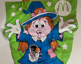 May include: A green plastic trick-or-treat bag with a witch character holding a spider. The bag has a yellow handle shaped like the McDonald's logo and a white McDonald's logo on the front.