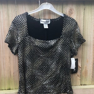 Puede incluir: Top negro de manga corta con cuello cuadrado y un patrón plateado en forma de red. La parte superior está colgada de una percha blanca contra una valla de madera. La etiqueta dice "Ronni Nicole".