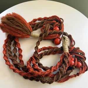 Könnte beinhalten: Ein langer, brauner und orangefarbener Makramee-Wandbehang mit Holzperlen. Der Behang hat oben einen Quasten.