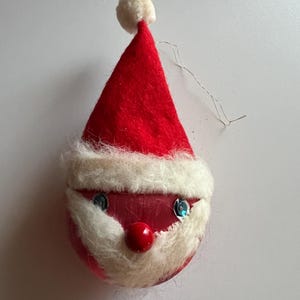 Puede incluir: Un adorno navideño vintage con forma de cara de Papá Noel. El adorno está hecho de fieltro rojo y tiene una barba y un gorro de piel blanca. Tiene ojos de purpurina azul y un pompón rojo en el gorro.