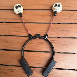 May include: A black headband with two white skull-shaped decorations. The skulls are connected to the headband with pink coiled springs. Rectangular black ear pieces are attached to the headband.