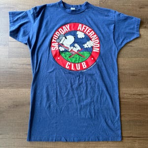 May include: A blue t-shirt featuring a circular graphic with the text "Saturday Afternoon Club." The graphic depicts a cartoon dog in a beach chair, with clouds and a red and green background.