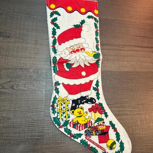 May include: A vintage Christmas stocking featuring a cartoon Santa Claus smoking a pipe. The stocking is white with a red top and is decorated with holly, gifts, and a yellow teddy bear. A festive holiday decoration.