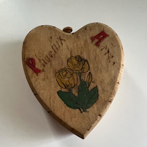 May include: A heart-shaped wooden box with a painted design of yellow roses and green leaves. The box is painted with the words "Phoenix Arts" in red and black.