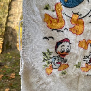 Op de afbeelding: Witte badstof handdoek met een cartoon eend ontwerp. De handdoek heeft een gefranjerde rand en is versierd met afbeeldingen van eenden in verschillende poses, met gele, rode, blauwe en groene accenten. De handdoek hangt buiten.