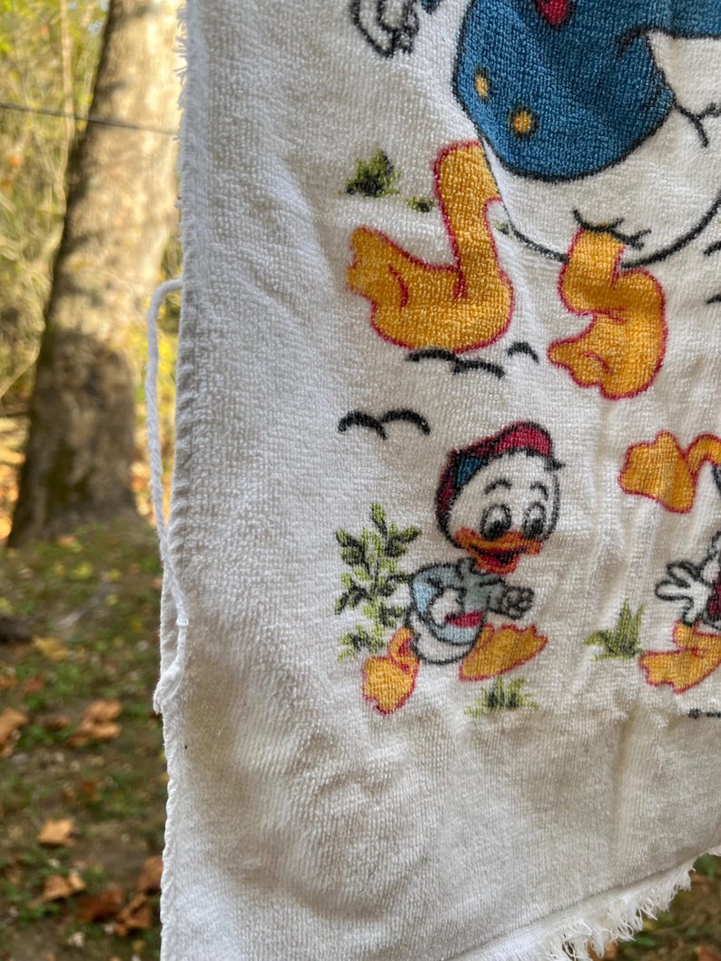 Op de afbeelding: Witte badstof handdoek met een cartoon eend ontwerp. De handdoek heeft een gefranjerde rand en is versierd met afbeeldingen van eenden in verschillende poses, met gele, rode, blauwe en groene accenten. De handdoek hangt buiten.