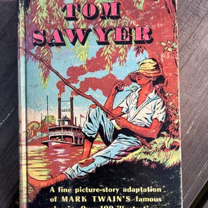 May include: A vintage paperback book cover featuring a colorful illustration of a boy fishing on a riverbank. The book title is "TOM SAWYER" in large, bold letters. The cover also includes text about the book being a picture-story adaptation of Mark Twain's classic with over 100 illustrations by Floyd Torbert.