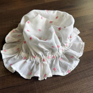 May include: White bonnet with pink embroidered rosebuds. The bonnet has a ruffled brim and elasticated neckline. The fabric is gathered at the neckline. The bonnet is on a dark wood surface.