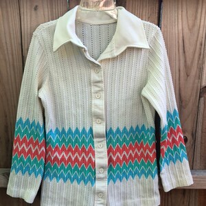 Op de afbeelding: Crèmekleurig vintage vest met knopen, een witte kraag en een zigzagpatroon in turquoise, rood en blauw op de mouwen en de onderkant. Het vest hangt aan een metalen hanger.