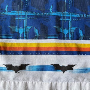 May include: A fabric with a Batman theme. The top portion features a dark blue pattern with light blue architectural designs and Batman symbols. Below is a horizontal stripe of yellow, orange, and blue. The bottom has the words "DARK KNIGHT" and Batman symbols.
