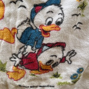 Op de afbeelding: Witte badstof handdoek met een cartoon illustratie van twee eenden. E&eacute;n eend draagt een blauwe jas, de andere een rode. De handdoek heeft een textuur en een grillig ontwerp, met vogels en bloemen.