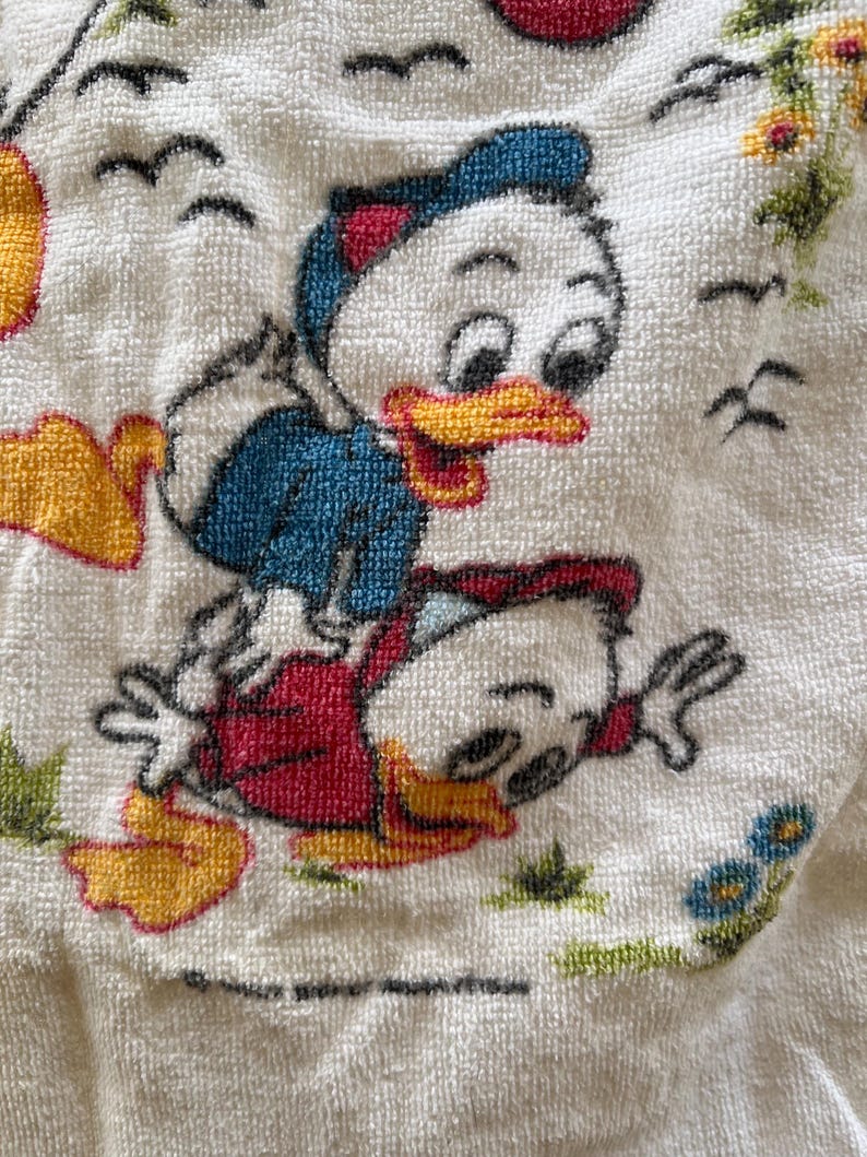 Op de afbeelding: Witte badstof handdoek met een cartoon illustratie van twee eenden. E&eacute;n eend draagt een blauwe jas, de andere een rode. De handdoek heeft een textuur en een grillig ontwerp, met vogels en bloemen.