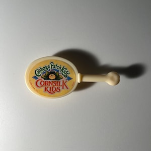 Puede incluir: Un juguete vintage de Cabbage Patch Kids Cornsilk Kids. El juguete es de color amarillo claro con una parte superior redonda y un asa. La parte superior presenta el logotipo de Cabbage Patch Kids y las palabras "Cornsilk Kids".