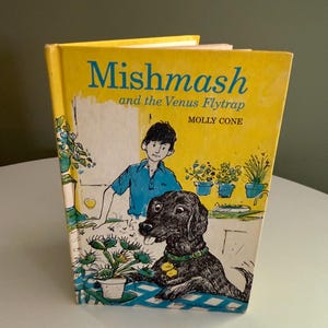 Może przedstawiać: Książka w twardej oprawie zatytułowana Mishmash and the Venus Flytrap autorstwa Molly Cone. Żółta okładka zawiera czarno-białą ilustrację osoby w niebieskiej koszuli, czarnego psa w zielonej obroży oraz muchołówki w doniczce na niebieskim stole.