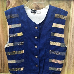 Puede incluir: Un chaleco azul sin mangas con forro de algodón blanco y cierre de botones. El chaleco está hecho de una tela de patchwork con diferentes patrones en tonos de marrón, beige y blanco. El chaleco tiene una etiqueta que dice "Nativewear Designs" y "M".