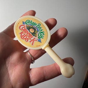 May include: A vintage Cabbage Patch Kids hand mirror. The oval mirror has a cream-colored handle and a yellow and white face with the words "Cabbage Patch Kids" and a cartoon image. The mirror is held in a hand.