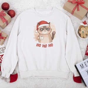 Może przedstawiać: Biała bluza z kapturem z wściekłym kotem w czapce Świętego Mikołaja, trzymającym kubek i napisem "HO HO HO". Prezenty świąteczne, ozdoby i skarpety otaczają bluzę.