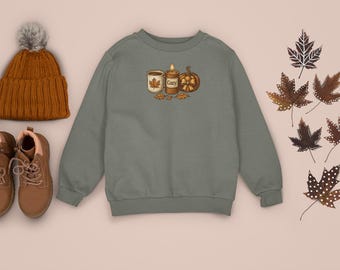 May include: An olive green sweatshirt with a fall-themed graphic, a brown knit beanie with a gray pom-pom, and brown boots with laces. Decorative fall leaves are scattered around the sweatshirt.