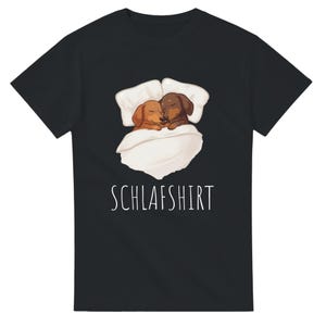 Może przedstawiać: Czarna koszulka z grafiką przedstawiającą dwa jamniki tulące się pod kocem i poduszkami. Biały napis "SCHLAFSHIRT" jest wydrukowany pod ilustracją. Psy są brązowe i płowe.