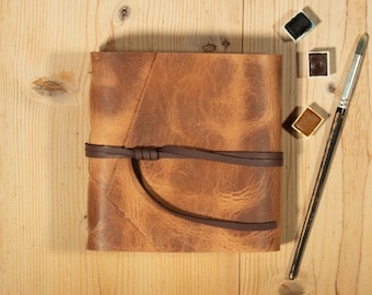 May include: A square, tan leather journal with a distressed texture, secured by a dark brown leather wrap-around tie. Beside the journal are three small, square watercolor paint pans in black, dark brown, and ochre, and a long, thin paintbrush.