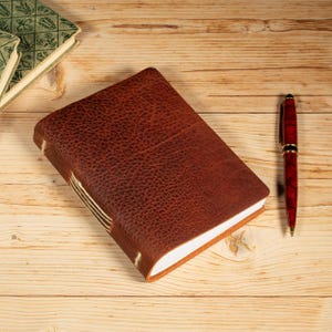 May include: A brown leather-bound journal with a textured surface and visible cream-colored stitching along the spine, featuring white pages. Beside the journal lies a red and black marbled ballpoint pen with gold-colored accents and a metal clip.