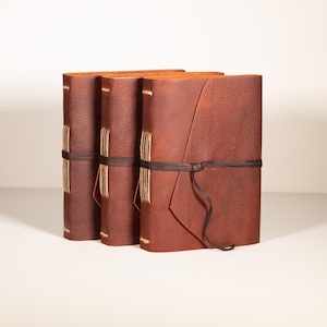 May include: Three brown leather journals with hand-stitched spines and wrap-around ties. The journals have a textured surface and a rustic aesthetic. The leather is a rich, warm brown color, and the stitching is a contrasting light tan.