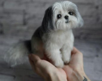 May include: A small, fluffy Shih Tzu dog figurine with a white and gray coat. The dog is sitting upright, with its front paws extended. The figurine has a sweet expression and is held in cupped hands.
