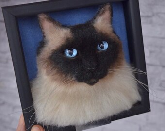 May include: Needle felted portrait of a Siamese cat with blue eyes in a black frame. The cat is light brown with a black face and a white chest.