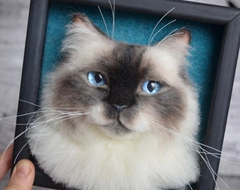 May include: A needle felted portrait of a white and gray cat with blue eyes, framed in a black frame with a teal blue background.