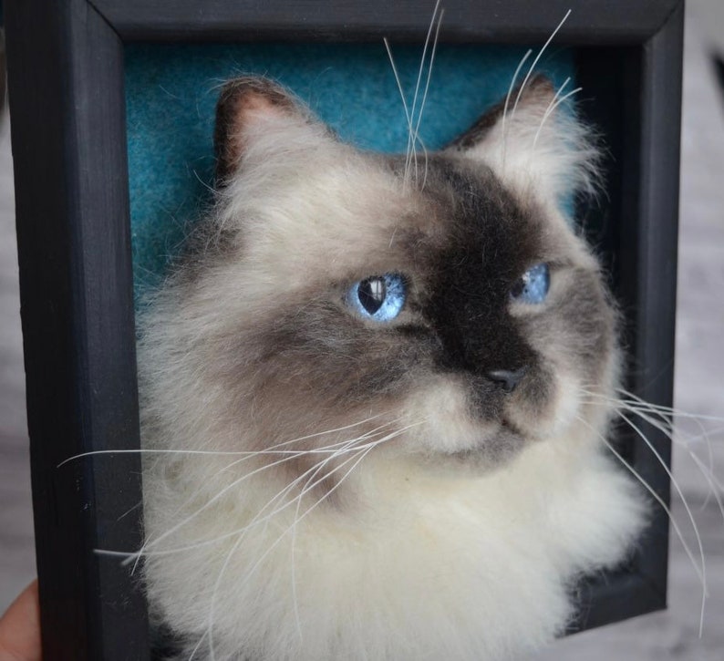 Pode incluir: Um close-up de um retrato de gato feito com feltro de agulha. O gato tem p&ecirc;lo branco com marcas cinzas e olhos azuis. O retrato est&aacute; emoldurado em uma moldura preta com um fundo azul turquesa.