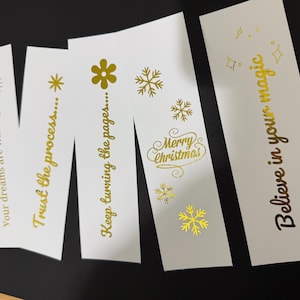 Può includere: Segnalibri di carta bianca con testo e disegni dorati. I segnalibri presentano frasi come "I tuoi sogni ti aspettano...", "Fidati del processo..." e "Credi nella tua magia". Un segnalibro dice "Buon Natale" con fiocchi di neve.